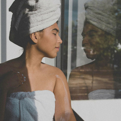 Woman in bath and head towel standing next to a window.