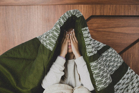 Woman sitting no bed with blankets covering her head and hands on her face.